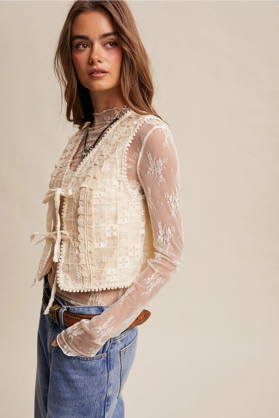 Woman wearing a sheer lace top with long sleeves and blue jeans on a beige background