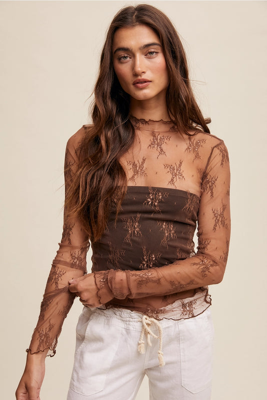 Woman wearing a sheer brown lace top with white pants on a beige background