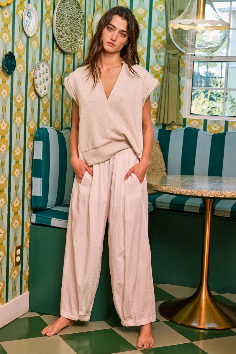 A woman wearing an oatmeal tucked sleeve sweatshirt with a deep V-neckline, cap sleeves, and contrast side details. The sweatshirt is on a casual outfit, paired with light-colored pants.